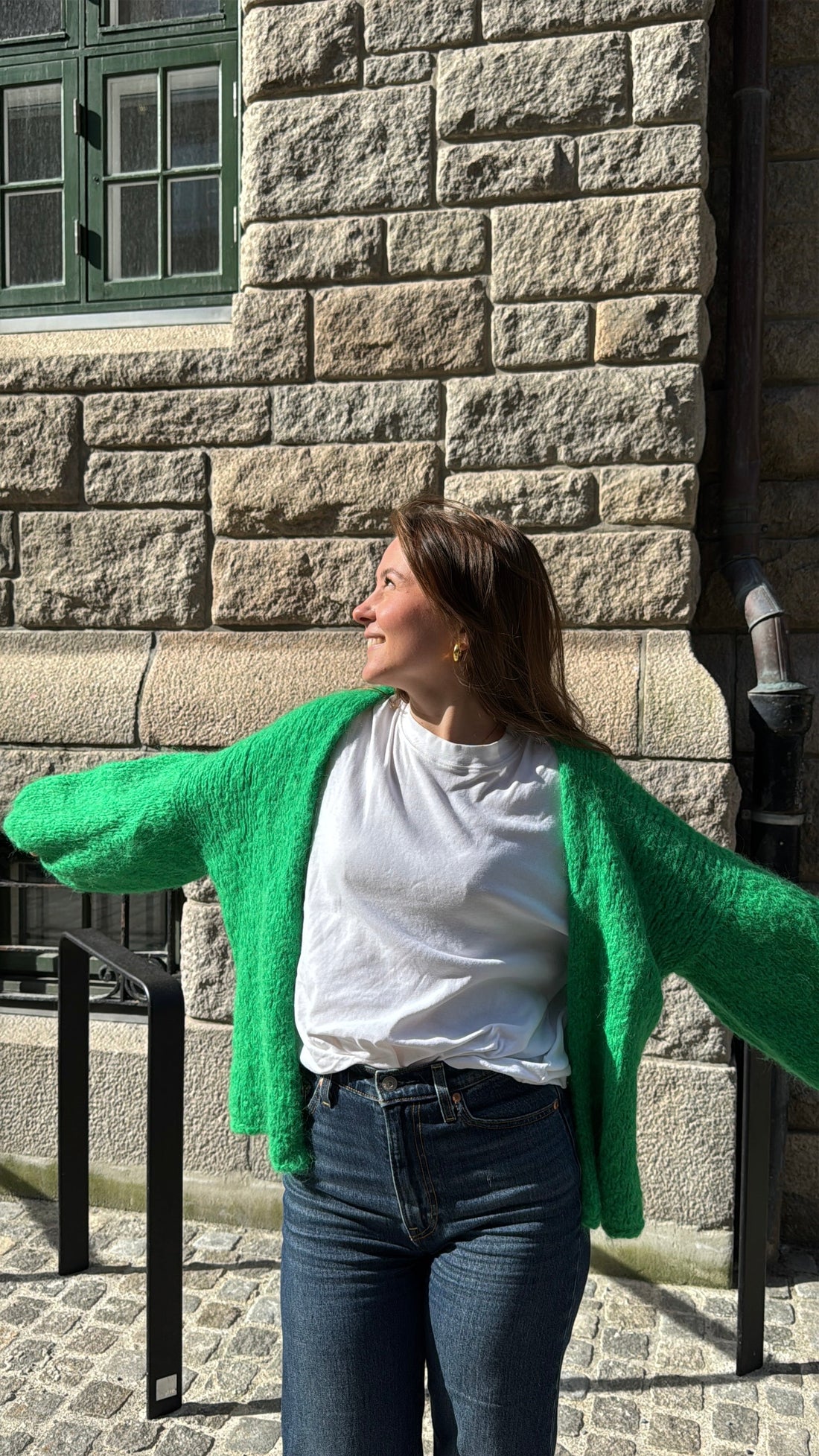 A knitter wearing a bright green hand knitted mohair cardigan over a white t-shirt in front of a stone wall