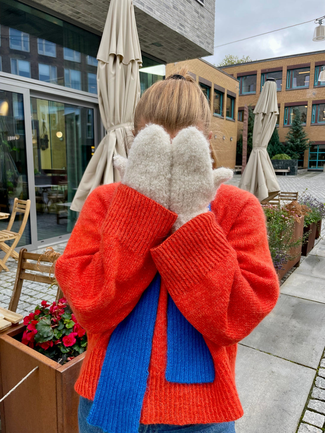 A knitter in a hand knitted orange sweater and blue scarf holding up a pair of cream knitted mittens