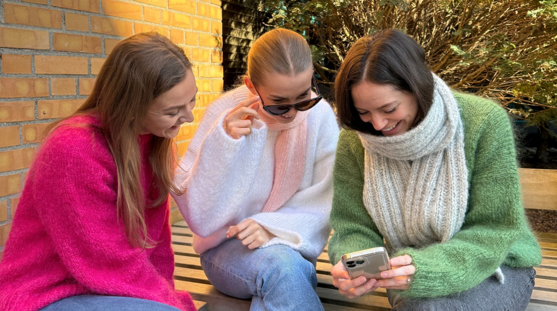 Three friends in hand knitted sweaters sitting together on a bench, looking at a knitting pattern on a phone