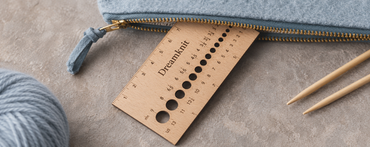Dreamknit wooden knitting needle gauge with US and millimeter size holes, resting on a zippered pouch beside a ball of yarn and circular knitting needles