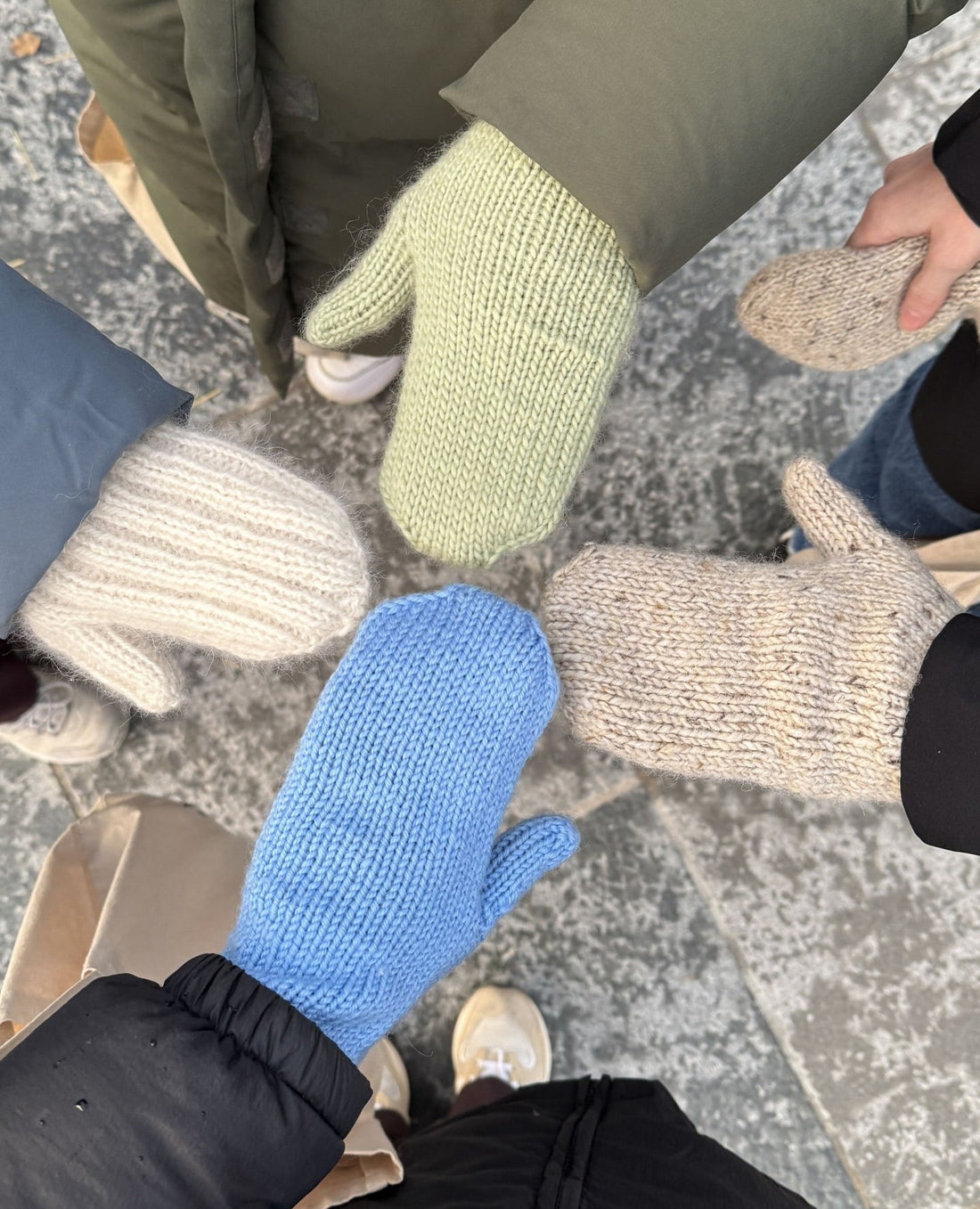 Four knitters holding their hands together in a circle wearing hand knitted mittens in green, blue, cream and beige tweed